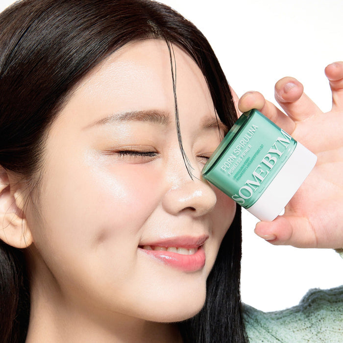 Woman holding SOME BY MI PDRN Spirulina Poreless Primer 10g next to her face, smiling with clear skin.