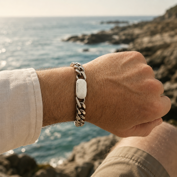 12mm stainless steel curb chain bracelet worn on the wrist overlooking a rocky coastal shoreline, showcasing the polished links and signature clasp in natural seaside light.